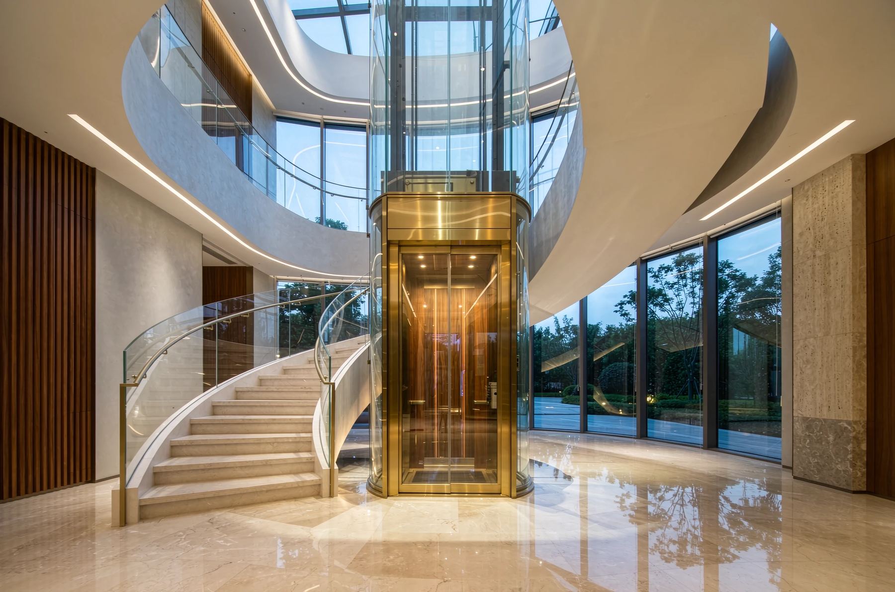 Glass-and-brass villa elevator integrated into a curved staircase in a double-height foyer